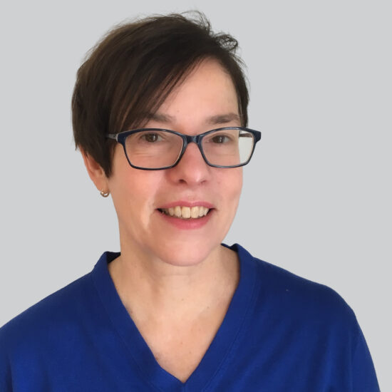 Buddhist Chaplain Linda Krause, a person with short brown hair and glasses, wearing a royal blue top, smiling against a plain light gray background.