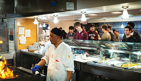 Jean Sauveur standing at a stove cooking with flames coming out a frying pan as a group of students watch.